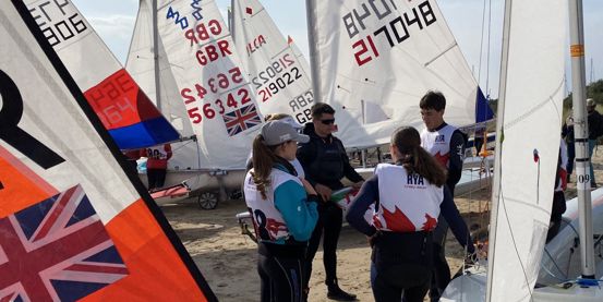 Young sailors chatting in a group alongside rigged boats on the beach at the Welsh Youth & Junior Championships, Plas Heli, 2025.
